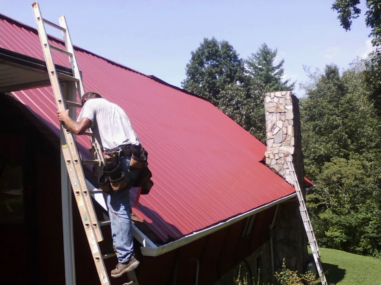 Roofer on a ladder beside a red metal roof and stone chimney — Soffit & Fascia Repair by Roofing Brothers in North Henderson